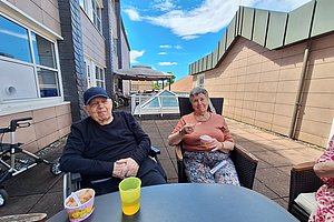 Zwei ältere Menschen sitzen auf einer Terrasse an einem Tisch mit Bechern und Schalen, im Hintergrund ein blauer Himmel mit Wolken und ein Gebäude
