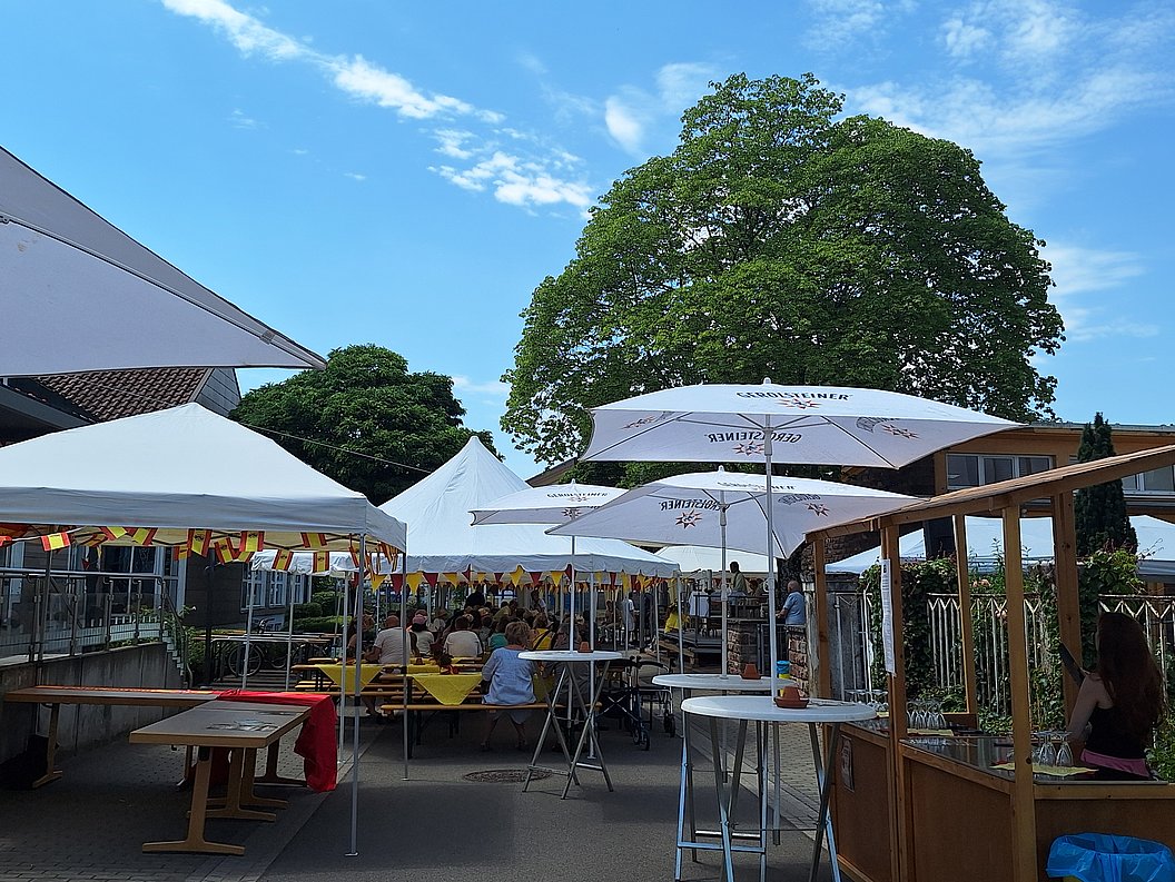 Sommerfest mit mehreren weißen Zelten, Sonnenschirmen und Sitzgruppen im Freien unter blauem Himmel und großen Bäumen