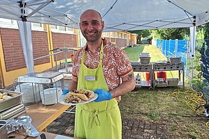 Mann mit grünem Schürze und blauen Handschuhen hält Teller mit Essen unter einem Pavillon im Freien