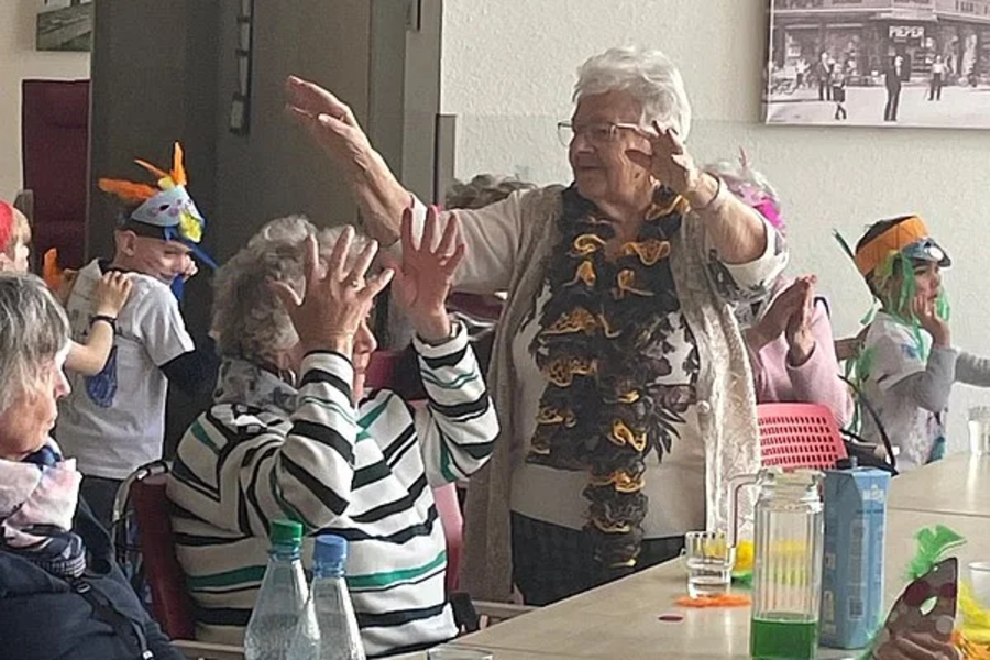 Mehrere ältere Menschen und Kinder sitzen an einem Tisch, einige tragen bunte Hüte, eine Person steht mit erhobenen Armen, bunte Papierdekoration hängt von der Decke.