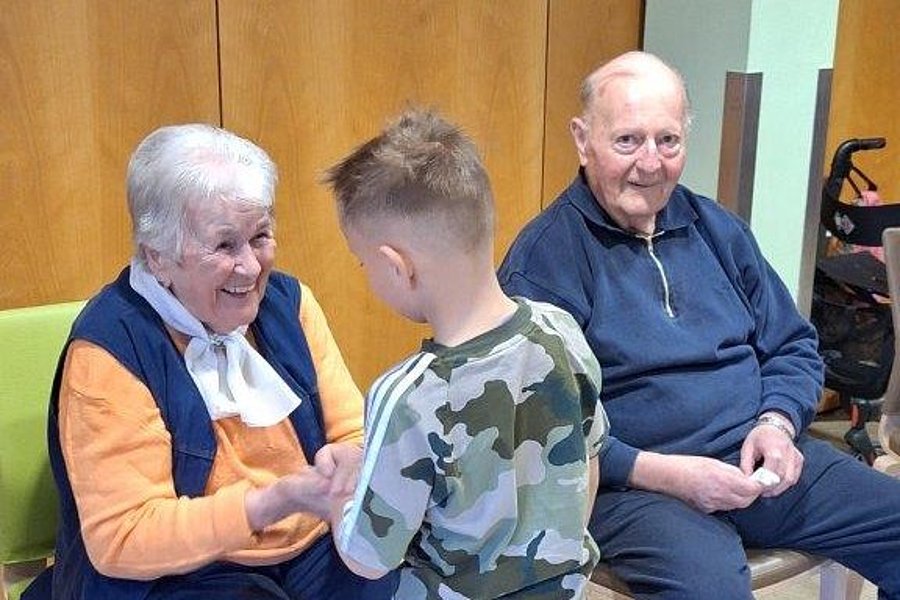 Junge in Camouflage-Pullover hält Hände einer älteren Frau mit orangefarbenem Pullover und weißem Halstuch, ein älterer Mann sitzt daneben auf Stuhl.
