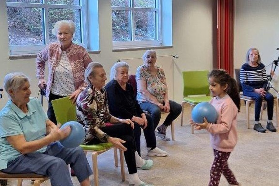 Mehrere ältere Menschen sitzen auf Stühlen in einem Raum, ein Kind hält einen blauen Ball und steht vor ihnen.