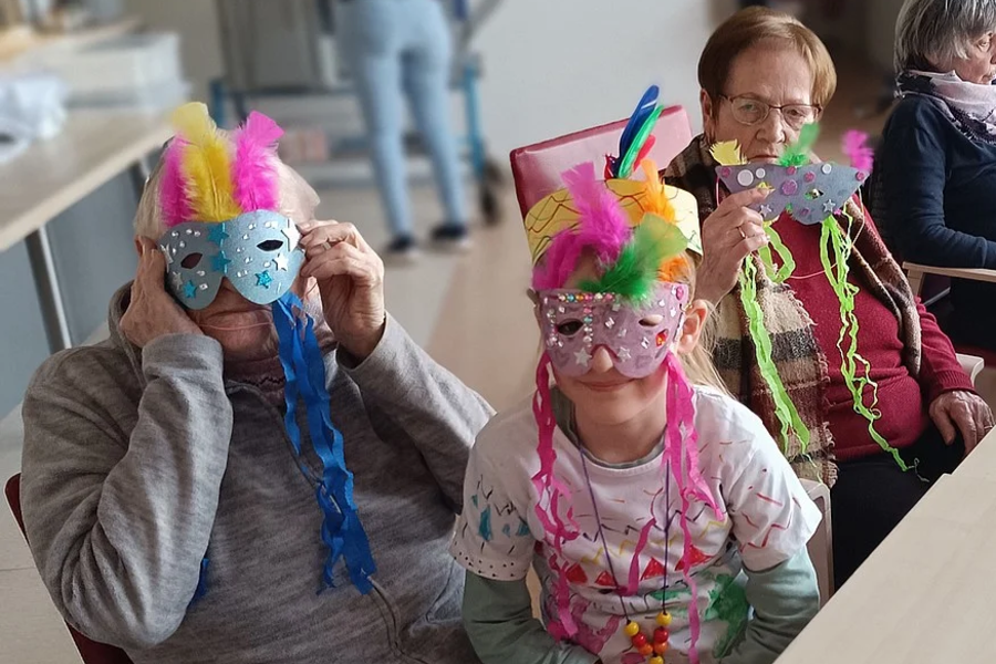 Drei Personen sitzen an einem Tisch, zwei tragen bunte Masken mit Federn und bunte Halsketten, eine trägt eine bunte Halskette.