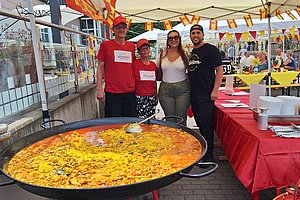 Vier Personen stehen hinter einer großen Pfanne mit Paella unter einem Pavillon mit spanischen Flaggen