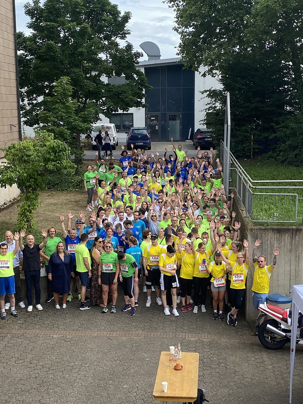 Große Gruppe von Menschen in bunten Sportshirts, viele heben die Arme, im Freien vor einem Gebäude mit Bäumen.