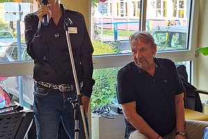 Zwei Männer in einem Raum mit großen Fenstern, einer steht mit Mikrofon vor einem Ständer, der andere sitzt auf einer Cajón, im Hintergrund Bäume und ein Gebäude sichtbar