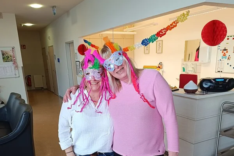 Zwei Frauen stehen in einem Flur, beide tragen bunte Partyhüte und eine trägt eine bunte Brille mit Federn.