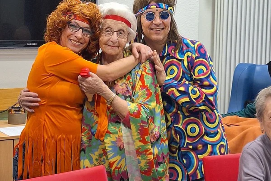 Drei Personen umarmen sich, eine trägt ein orangefarbenes Kleid, eine ein buntes Kleid mit Blumenmuster, die dritte ein buntes Hemd mit Kreis- und Wellenmustern.