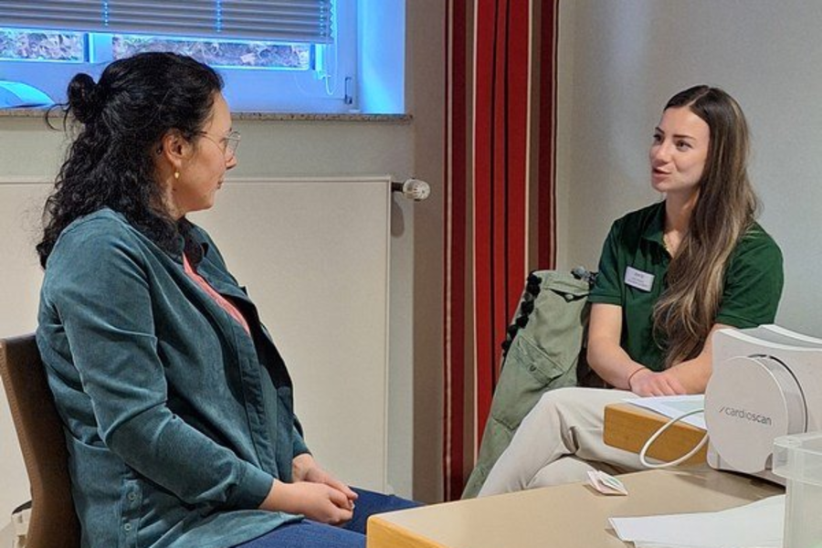 Zwei Frauen sitzen sich in einem Raum gegenüber, eine trägt eine grüne Jacke, die andere ein grünes Oberteil mit Namensschild.