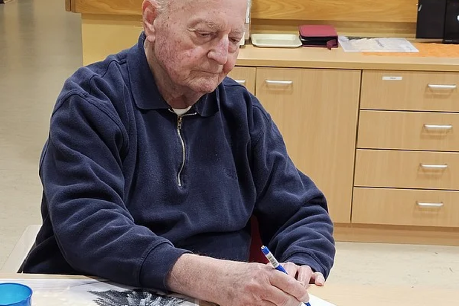 Älterer Mann in blauem Pullover sitzt an einem Tisch und schreibt auf einem Notizblock, auf dem Tisch liegen eine Zeitung und ein gefaltetes Papier.