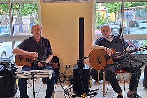 Zwei Männer sitzen nebeneinander und spielen akustische Gitarren vor einem großen Fenster mit Blick auf eine Straße und parkende Autos