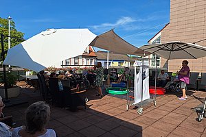 Gruppe älterer Menschen sitzt im Freien auf Terrasse unter Sonnenschirmen und Sonnensegeln