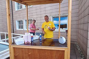Zwei Personen stehen hinter einem kleinen Holzverkaufsstand mit Dach auf einem gepflasterten Hof vor einem mehrstöckigen Gebäude