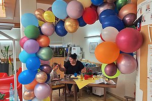 Bunte Luftballons bilden einen Bogen über einem Tisch, an dem eine Frau und ein Kind sitzen und sich gegenüberstehen