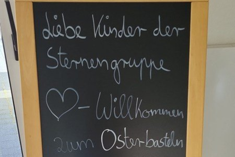 Holztafel mit schwarzer Schreibfläche, darauf weiße Kreideaufschrift: 'Liebe Kinder der Sternengruppe ♥-Willkommen zum Osterbasteln in der Tagespflege' und eine gezeichnete Hasenfigur.
