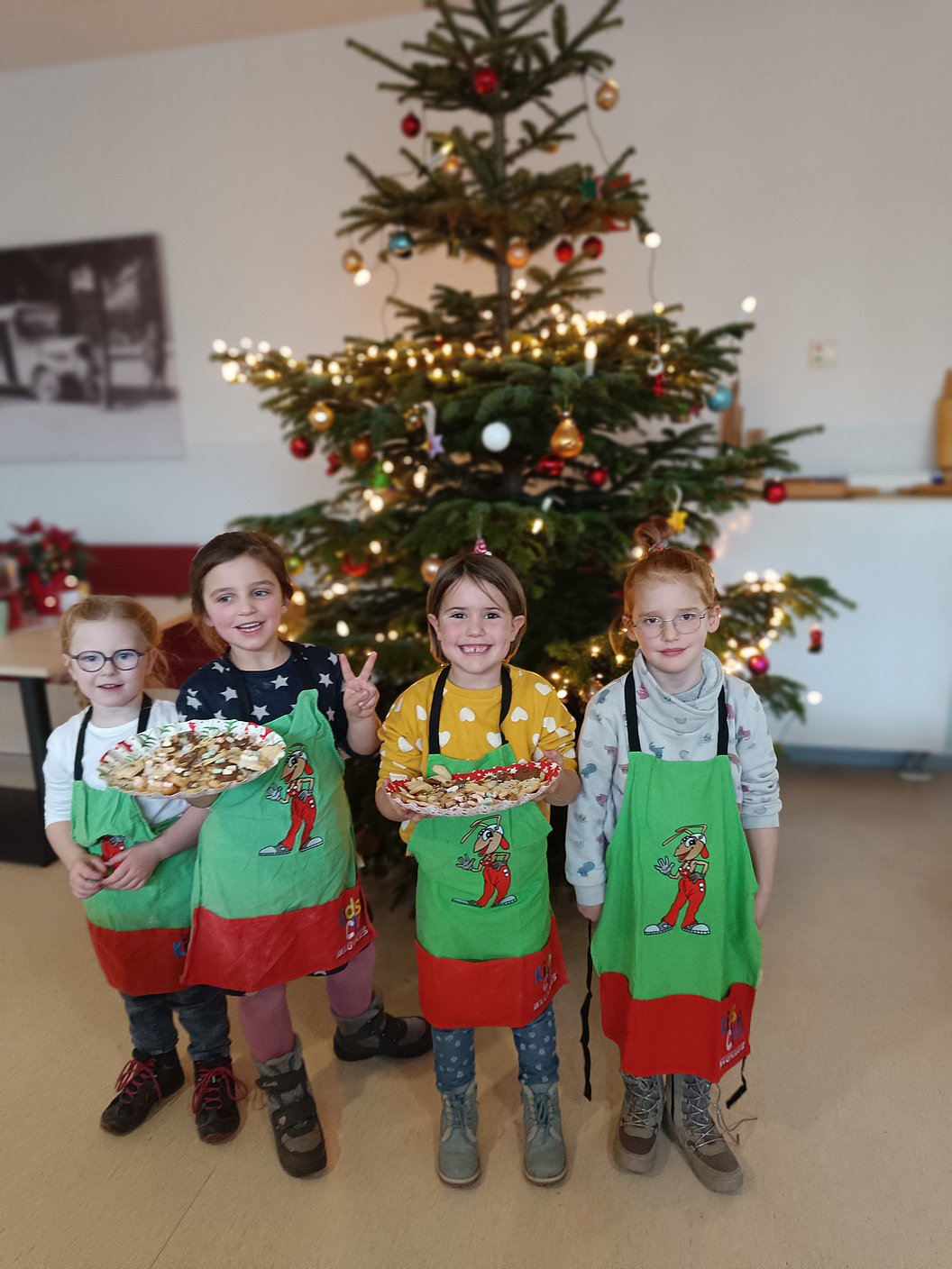 Vier Kinder mit grünen Schürzen und roten Taschen halten Platten mit Gebäck vor einem geschmückten Weihnachtsbaum.
