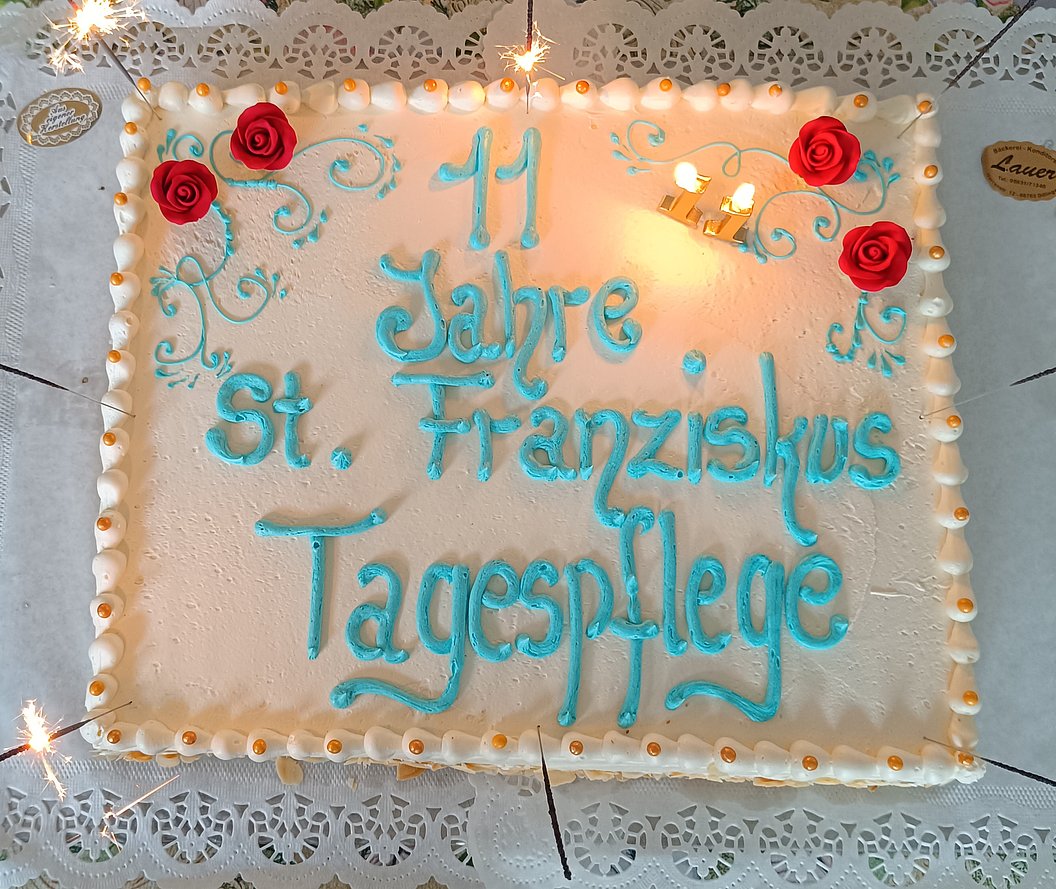 Rechteckige Torte mit blauer Schrift '11 Jahre St. Franziskus Tagespflege', verziert mit roten Rosen und Zuckergussrand, brennende Kerzen.