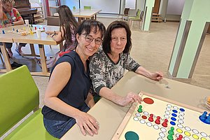 Zwei Personen sitzen an einem Tisch und spielen ein Brettspiel mit bunten Spielfiguren auf einem quadratischen Spielbrett