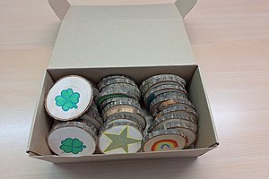 Offene Pappschachtel mit mehreren Holzscheiben, die mit bunten Symbolen wie Kleeblatt, Stern und Regenbogen bemalt sind