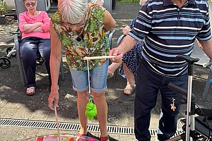 Ältere Frau und älterer Mann spielen mit Angelruten an einem kleinen Planschbecken im Freien, im Hintergrund sitzt eine weitere ältere Frau auf einem Stuhl.