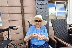 Ältere Frau mit Sonnenhut und Sonnenbrille sitzt an einem Tisch im Freien und hält ein Glas