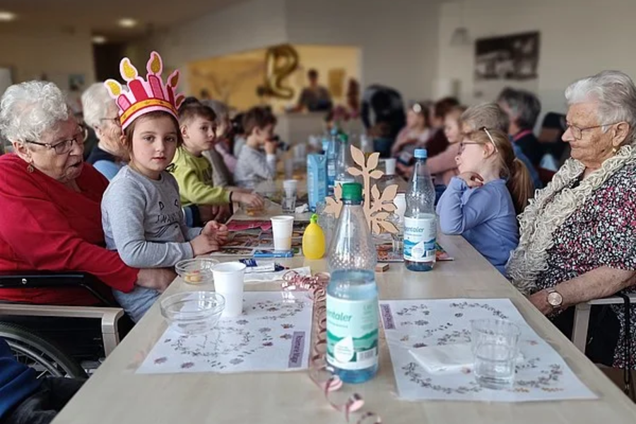 Mehrere Personen unterschiedlichen Alters sitzen an einem langen Tisch mit Getränkeflaschen und Papieren, ein Kind trägt eine bunte Papierkrone.