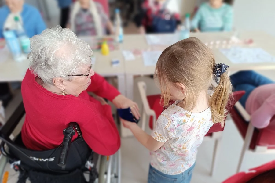 Ein Mädchen mit Pferdeschwanz hält die Hand einer älteren Person im Rollstuhl, im Hintergrund sitzen weitere ältere Menschen an einem Tisch.