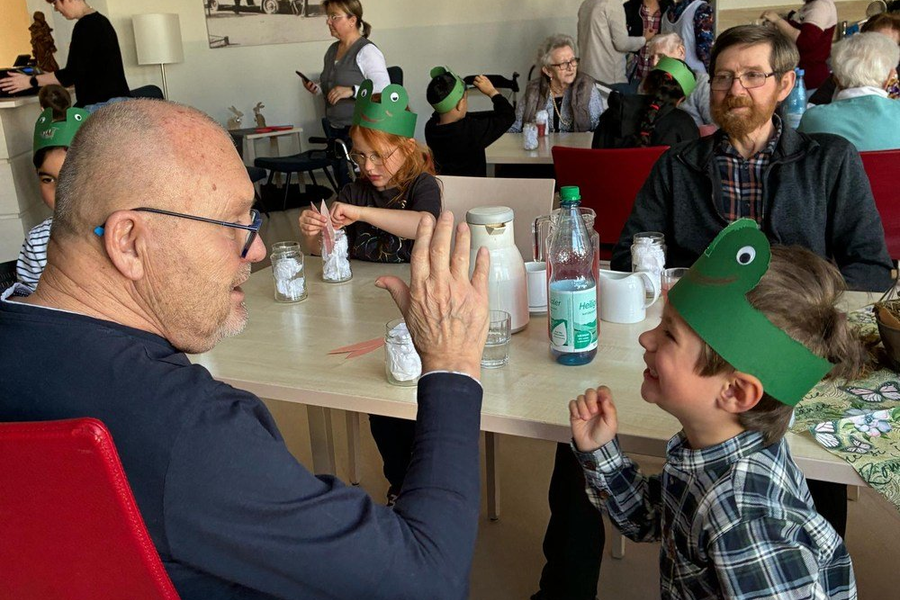 Mehrere Personen unterschiedlichen Alters sitzen an Tischen, tragen grüne Papiertierkronen und halten kleine weiße Objekte in den Händen.