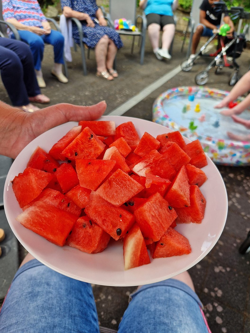 Nahaufnahme eines weißen Tellers mit gewürfelter Wassermelone vor einer Gruppe sitzender Menschen und einem Planschbecken mit Spielzeug im Hintergrund