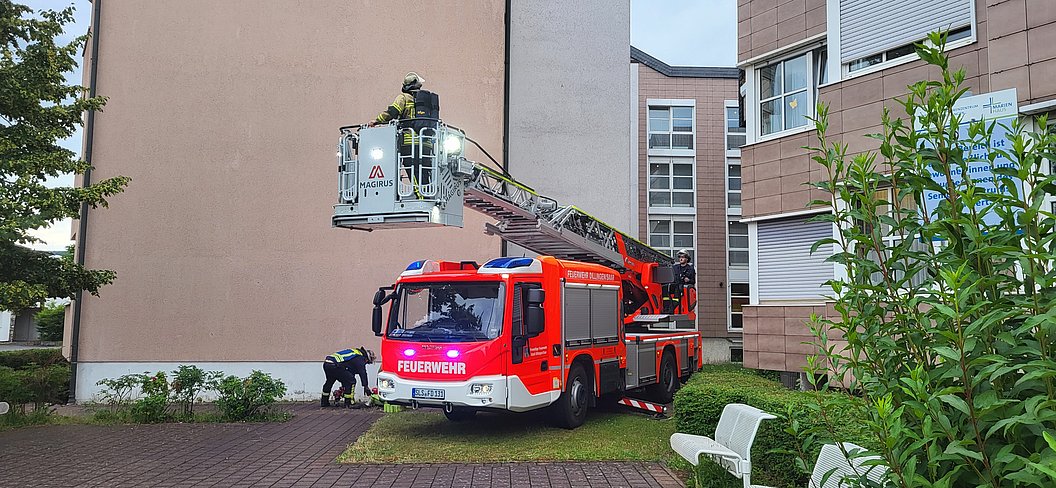 Rotes Feuerwehrfahrzeug mit ausgefahrener Drehleiter vor einem mehrstöckigen Wohngebäude, Feuerwehrleute auf der Leiter und am Boden.