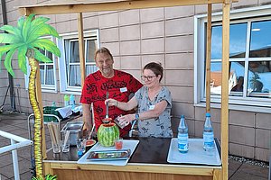 Zwei Personen an einem kleinen Holzstand mit einer Wassermelone und zwei Wasserflaschen auf der Theke vor einer hellen Hauswand mit Fenstern