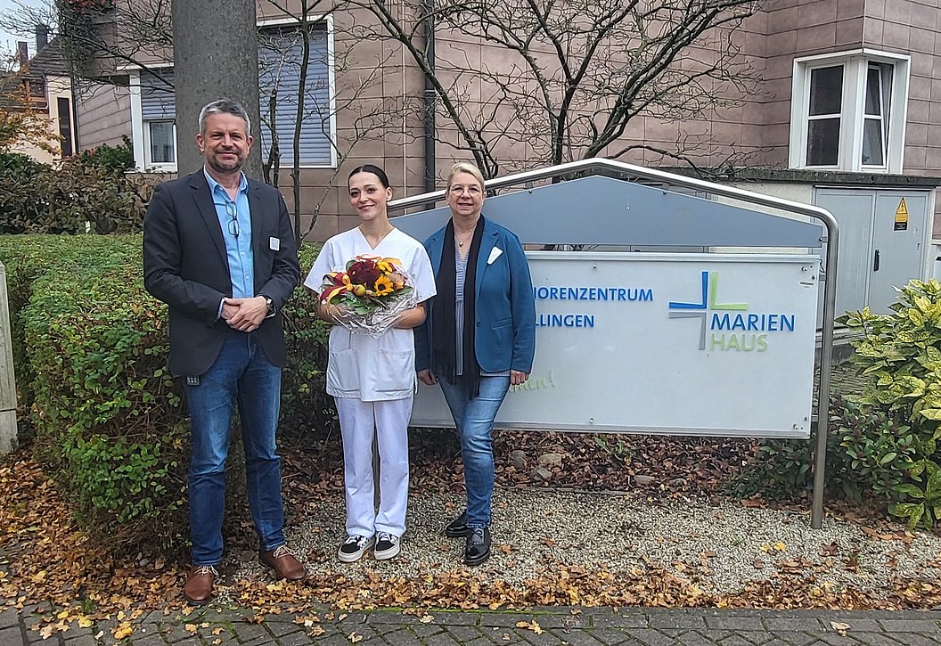 Drei Personen stehen vor einem Schild, eine trägt weiße Arbeitskleidung und hält einen Blumenstrauß.