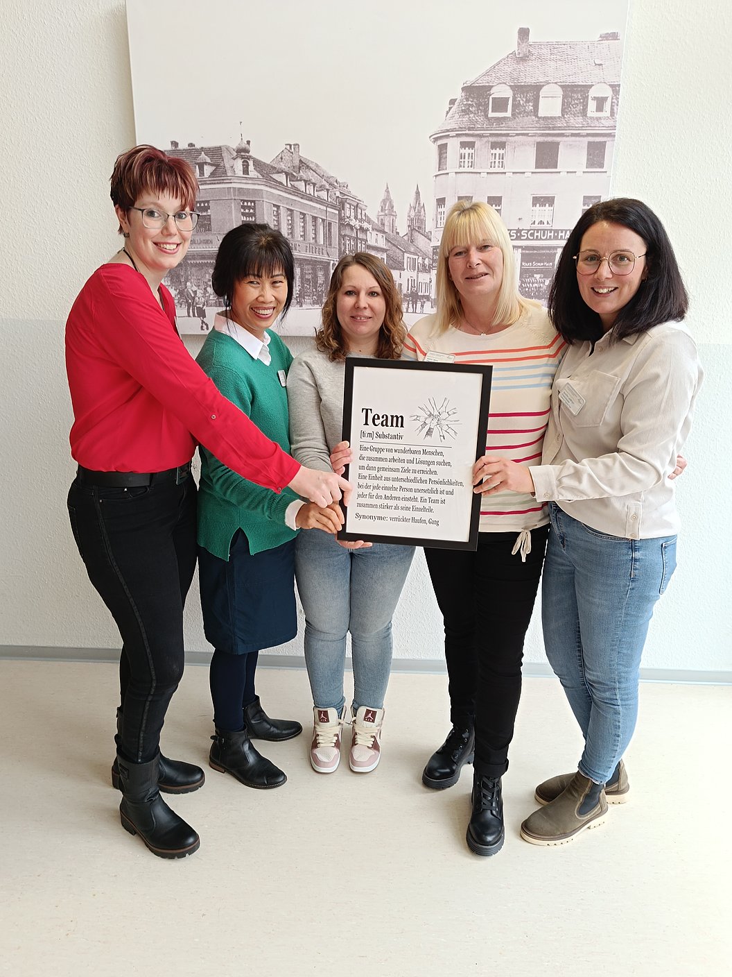 Fünf Frauen stehen in einem Raum vor einer schwarz-weißen Stadtbild-Wandgrafik und halten gemeinsam einen gerahmten Text mit der Überschrift 'Team'.