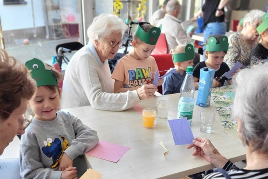 Mehrere ältere Menschen und Kinder sitzen an einem Tisch, schneiden buntes Papier und tragen grüne Papierkronen.