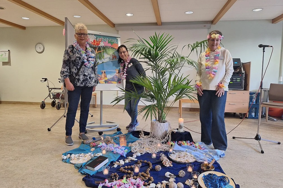 Drei Personen stehen um eine Bodenfläche mit blauer Decke, Muscheln, Blumenketten und einer kleinen Palme in einem Raum mit Holzdecke und Rollator.
