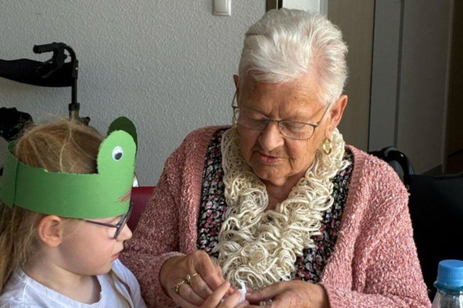 Ältere Frau und Kind sitzen an einem Tisch mit zwei leeren Gläsern und einer Wasserflasche, das Kind trägt ein grünes Tierkopfband.