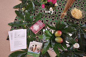 Auf einem gemusterten Tuch liegen große grüne Blätter, zwei Mangos, ein Besen, eine Schale mit kleinen gelben Stücken, zwei Bücher und mehrere lose Blätter mit Text.