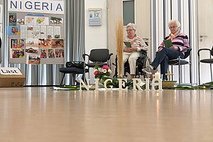 Zwei ältere Personen sitzen auf Stühlen und lesen vor einer Wand mit einer großen Tafel mit Fotos und dem Schriftzug 'NIGERIA'