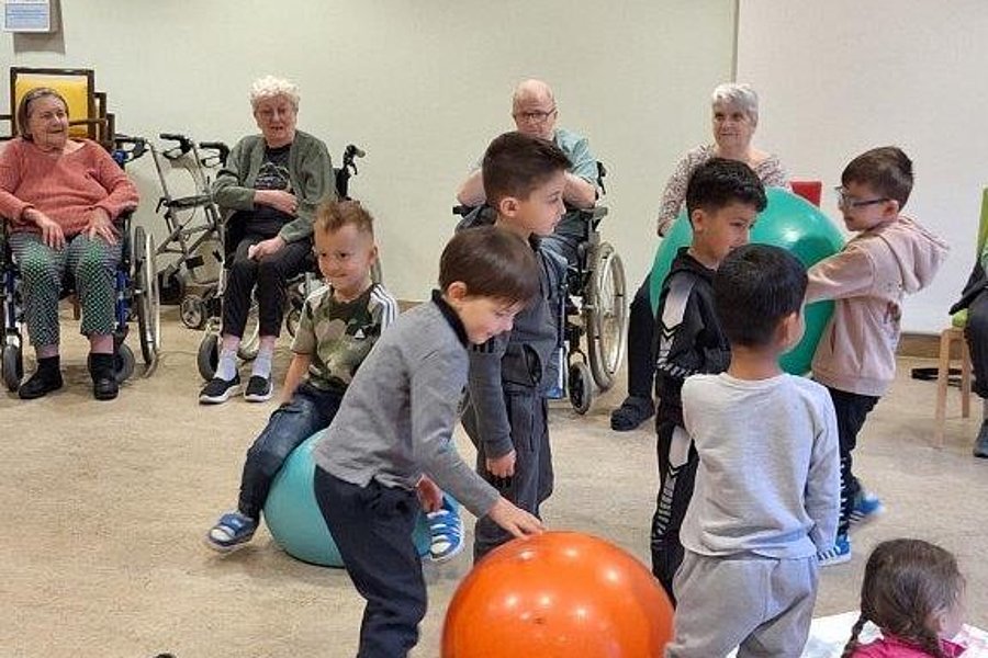 Mehrere Kinder spielen mit einem großen orangen Ball auf einer Decke in einem Raum, im Hintergrund sitzen ältere Menschen in Rollstühlen und Stühlen.
