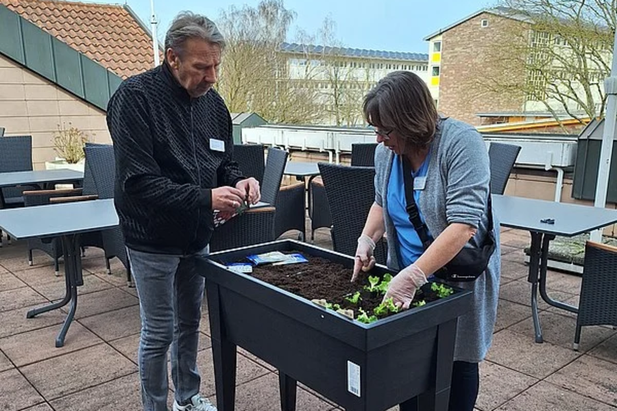 Zwei Personen stehen an einem schwarzen Hochbeet auf einer Terrasse, eine Person pflanzt kleine grüne Pflanzen ein.