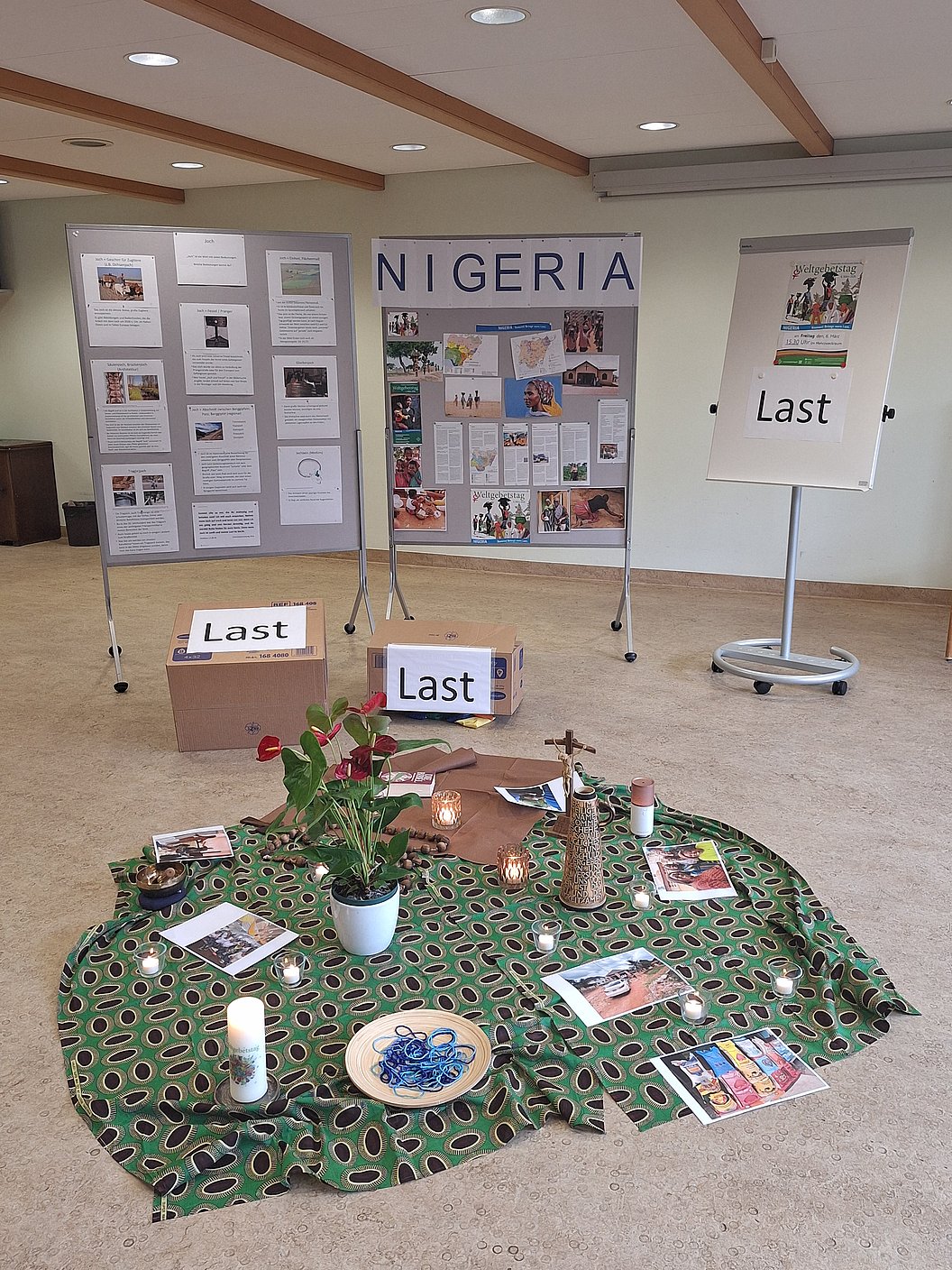 Ausstellung mit drei Stellwänden, auf denen Informationen und Bilder zu Nigeria und dem Thema "Last" gezeigt werden, davor ein runder Tisch mit grün gemustertem Tuch, Pflanzen, Kerzen und Büchern.