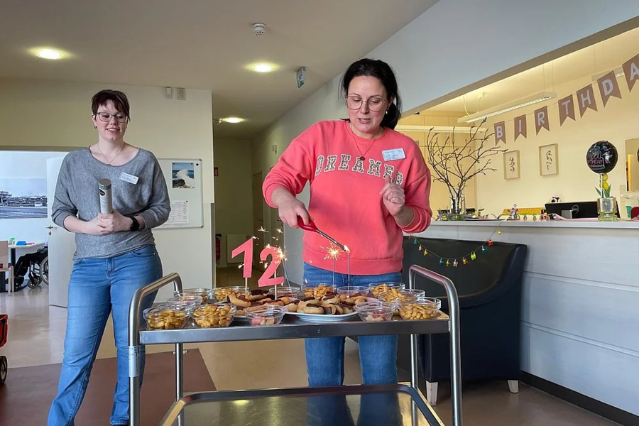 Zwei Frauen stehen neben einem Servierwagen mit verschiedenen Snacks und einer roten Zahl '12' aus Kerzen darauf.