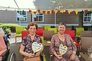 Zwei ältere Frauen sitzen unter einem weißen Zelt auf bunten Stühlen und halten herzförmige Schilder mit der Aufschrift 'Ilke St. Franziskus'.