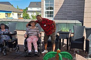 Mann beugt sich zu Frau im Rollstuhl auf Terrasse mit Gartenmöbeln und Planschbecken mit Spielzeug.