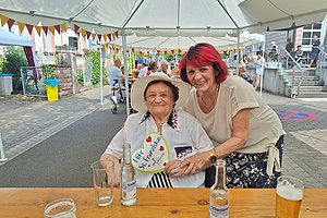 Zwei Frauen sitzen an einem Holztisch im Freien, eine ältere mit Hut hält ein Herz mit der Aufschrift 'I like St. Franziskus', die andere umarmt sie von der Seite, im Hintergrund bunte Wimpel und weitere Personen unter einem Zelt