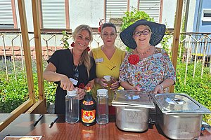 Drei Frauen hinter einem Holztisch mit Getränkekaraffen, einer Flasche und zwei Edelstahlbehältern, eine Frau trägt einen großen Hut und eine bunte Bluse