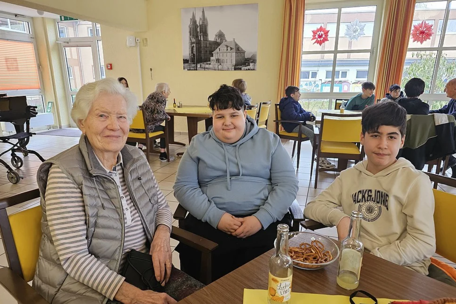 Drei Personen sitzen an einem Tisch in einem hellen Raum mit großen Fenstern und mehreren weiteren Tischen und Stühlen.