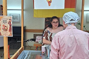 Verkaufsstand mit Schild 'Eisbude' und Eiscremeabbildung, davor zwei Personen, eine mit Kochmütze und Schürze, im Hintergrund deutsche Fahnen