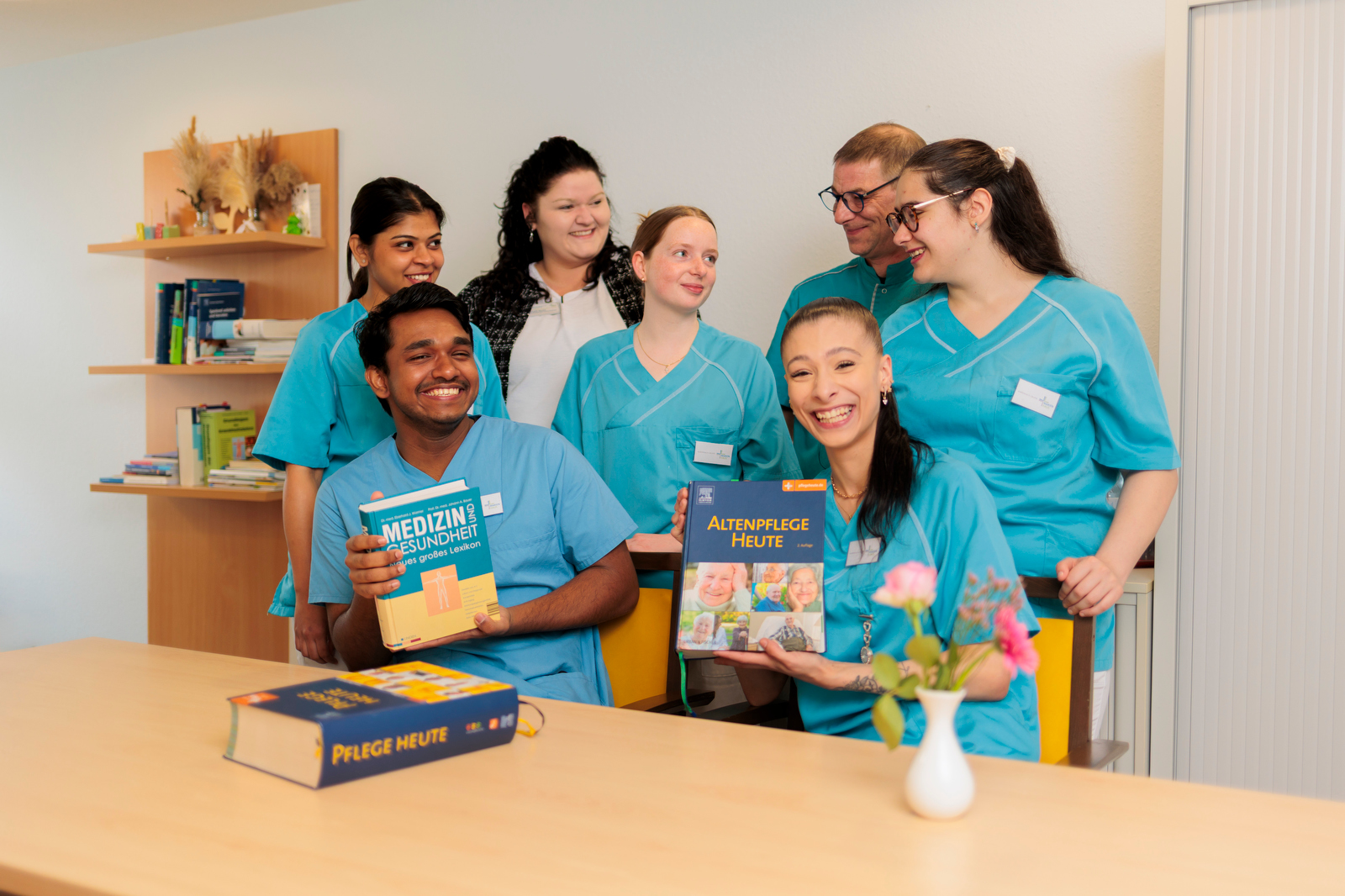 Gruppe von sechs Pflegekräften in blauen Kitteln, zwei halten Fachbücher, sie sitzen und stehen um einen Tisch mit einer kleinen Vase und Blumen