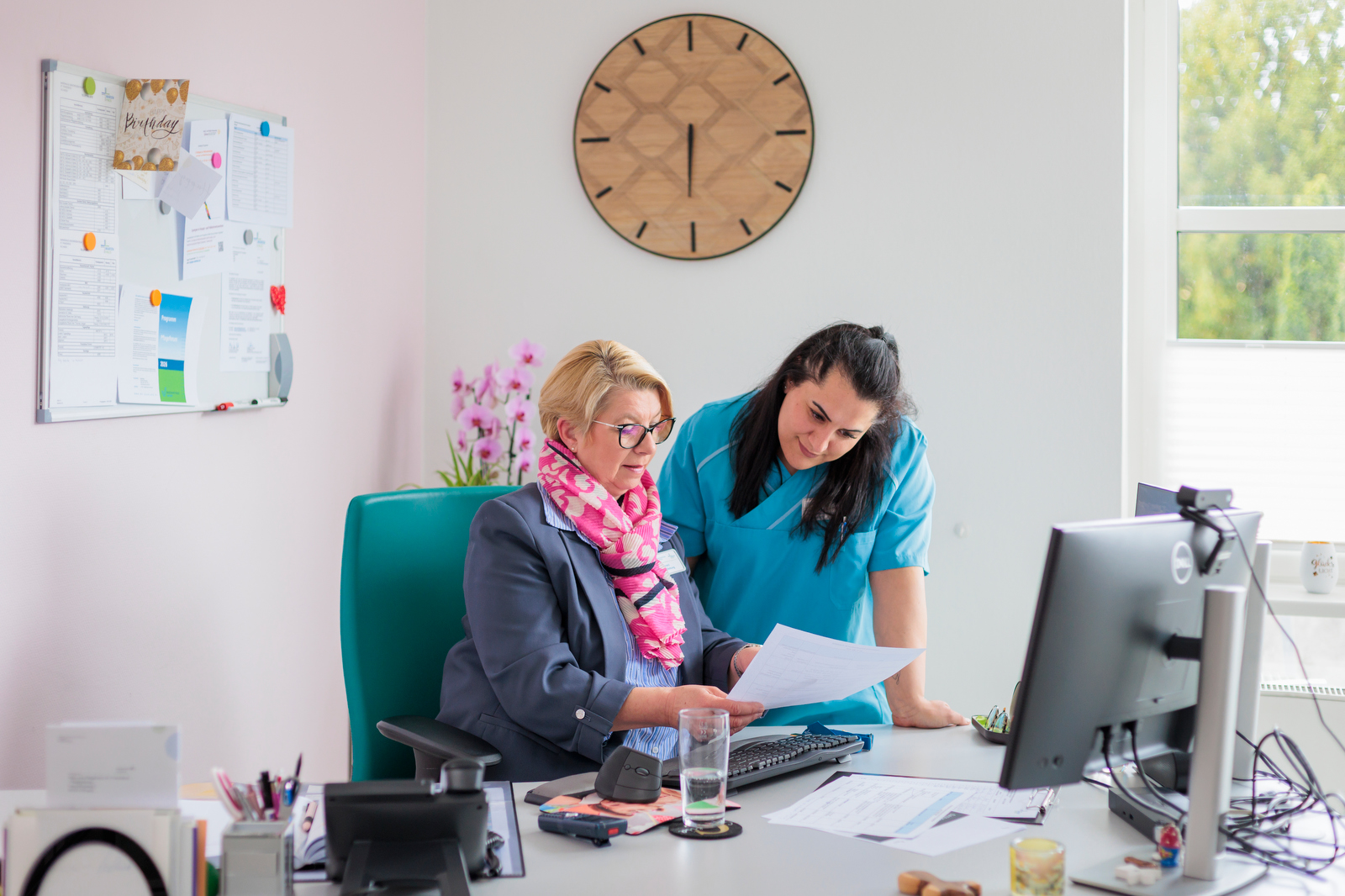 Zwei Frauen im Büro, eine sitzt am Schreibtisch und zeigt der anderen ein Dokument, im Hintergrund eine große Wanduhr und ein Fenster